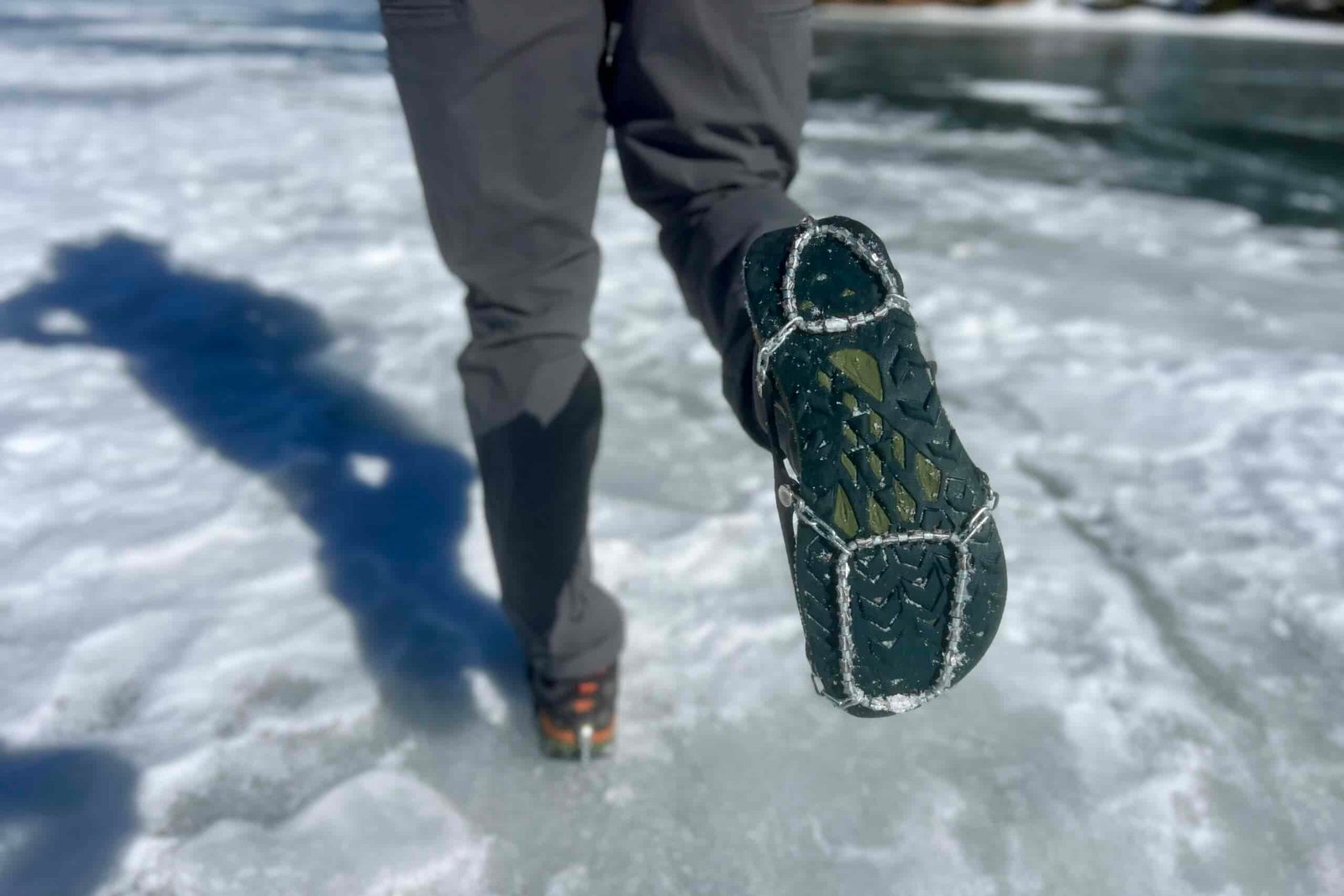 A hiker stands on a frozen lake and shoes the bottom of his shoe which is wearing an ice cleat.