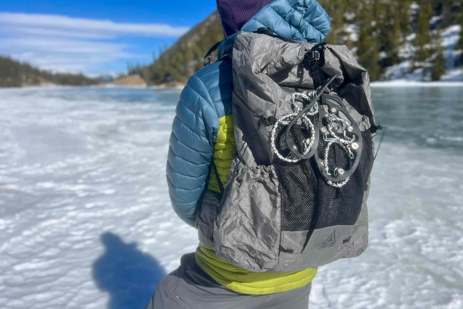 A hiker looks away from the camera on a frozen lake while wearing a backpack with ice cleats attached to the outside.