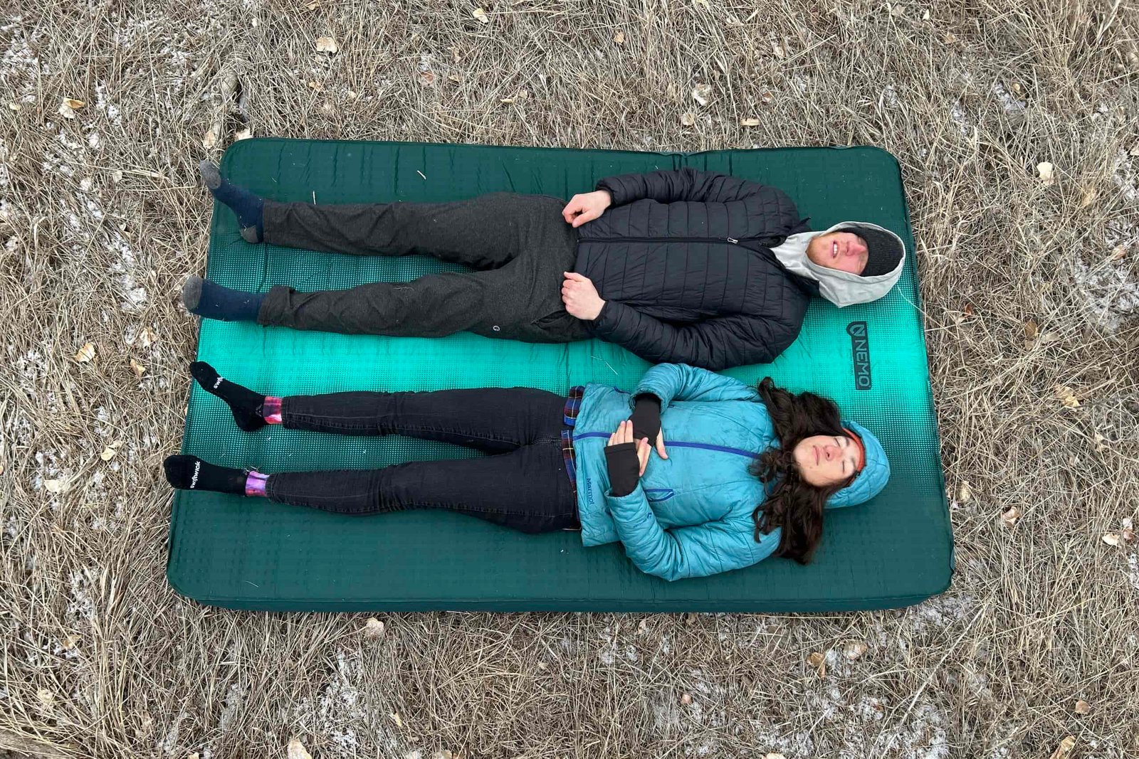 Top-down image of two people resting on a camping mattress on the grass.