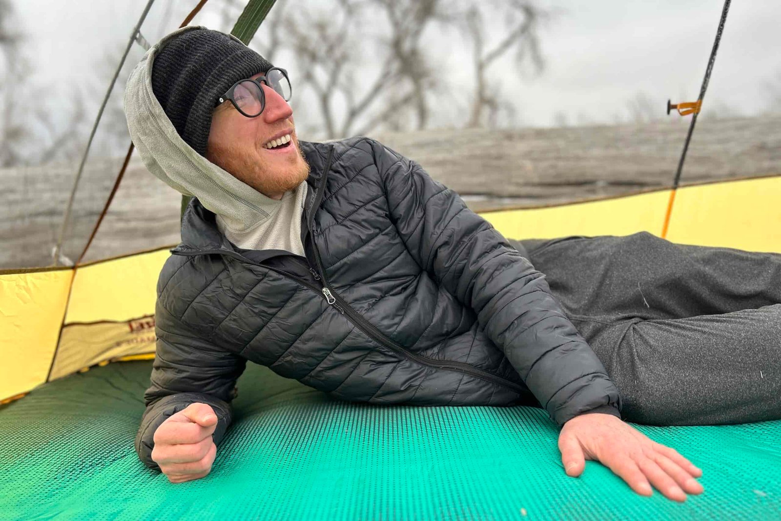 Close-up image of a man laying on a camping mattress inside a tent. He is smiling and looking off in the distance.