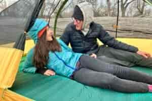 A woman and man are smiling and talking while laying on a camping mattress in a tent in the woods.
