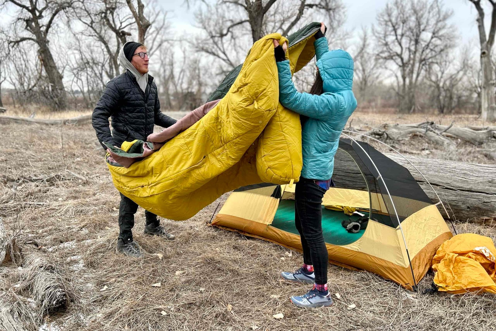 Two people fluff a double sleeping bag outside of a tent in a wintry forest setting.