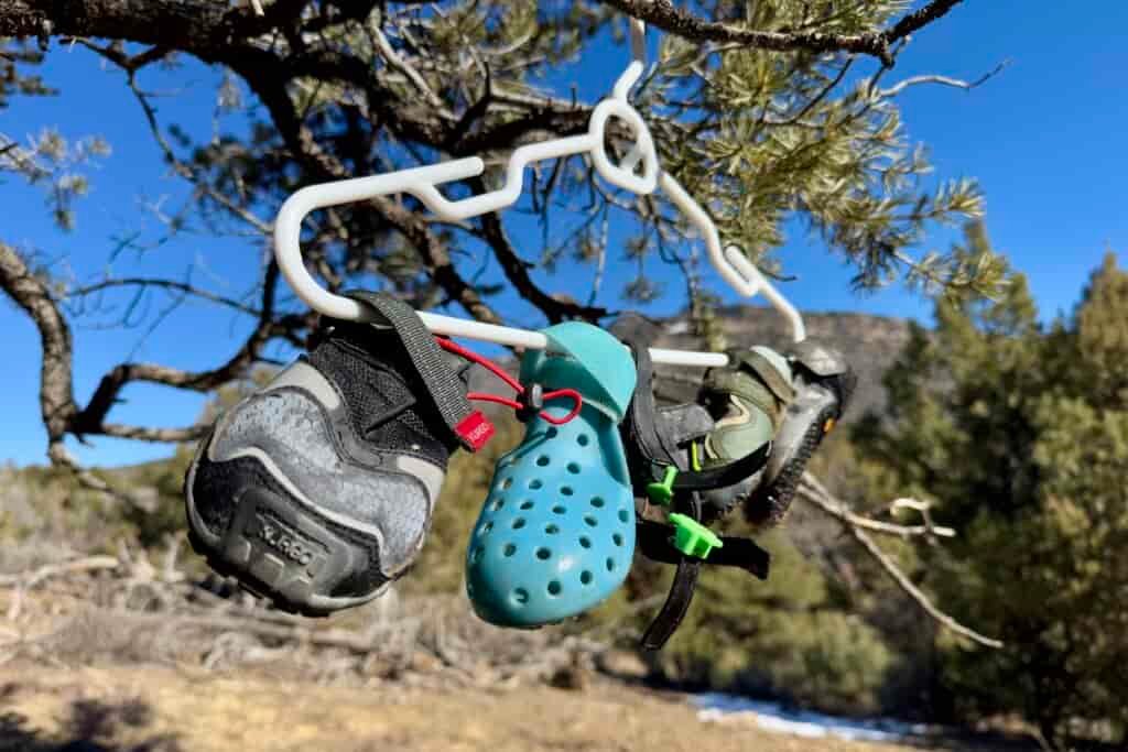 Several dog boots are attached to a clothes hanger by their velcro. The hanger is on a tree branch in the sunshine out in the woods.