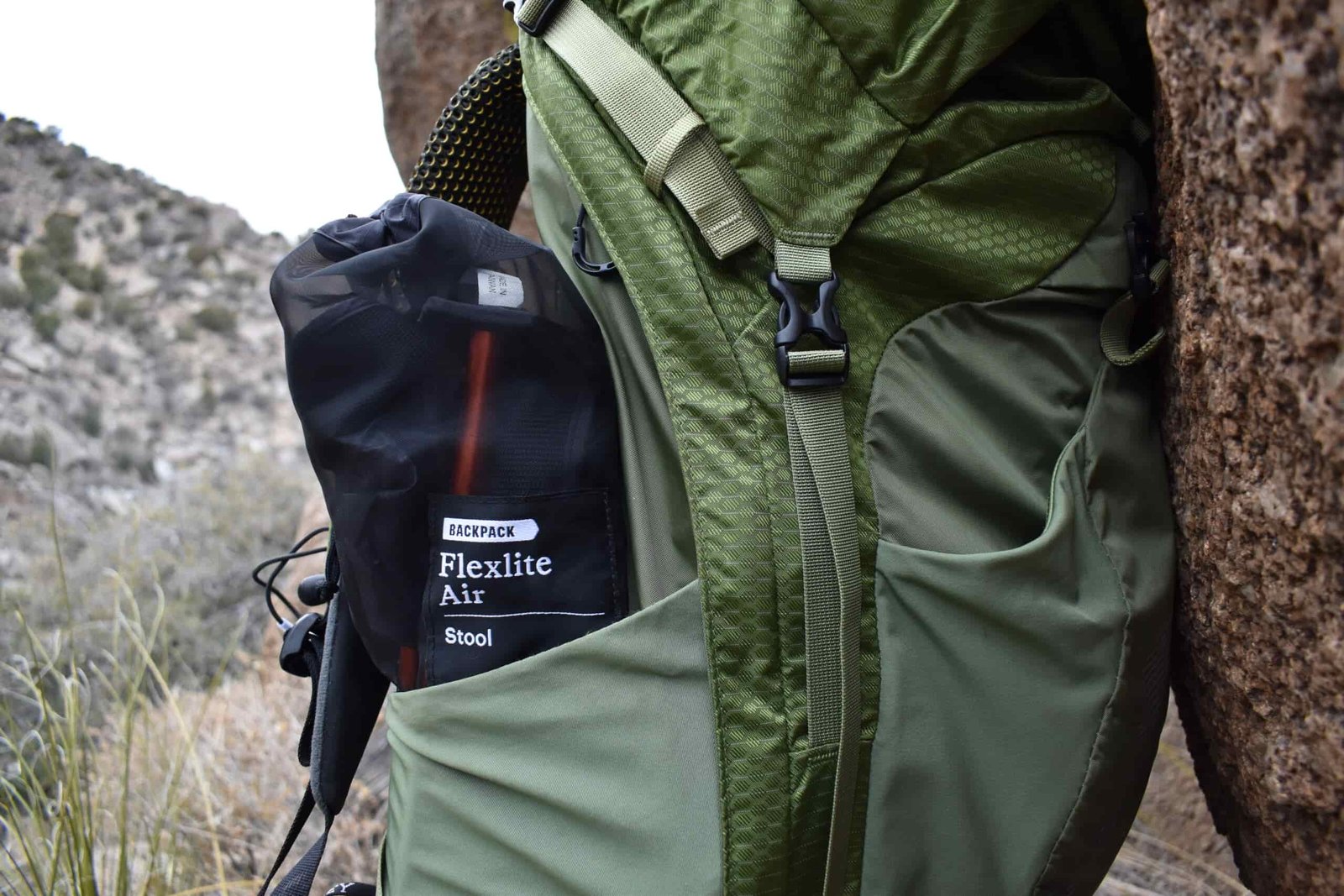The stuff sack of the REI stool sticks out of the side pocket of a green backpack
