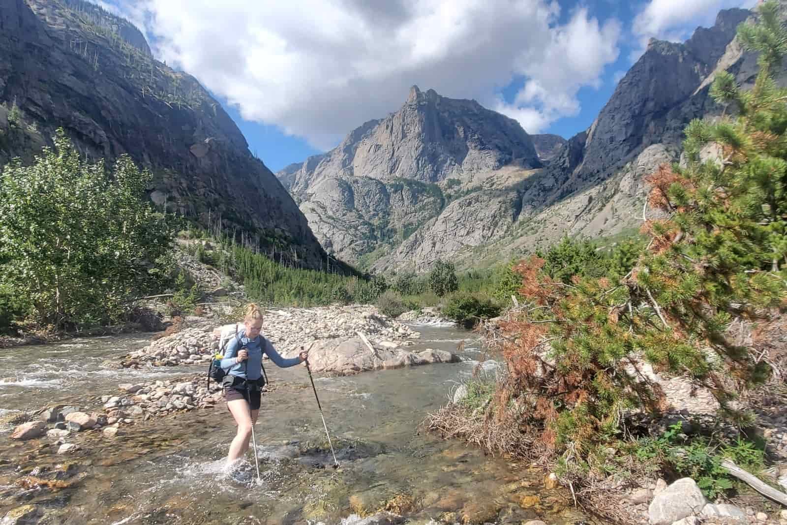 A woman crosses a river with mountain views.