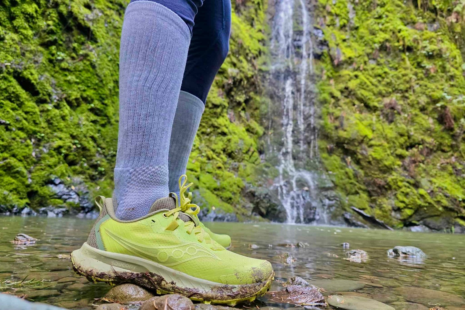 Hiker standing in front of a waterfall, with a focus on the Saucony shoes that she is wearing.