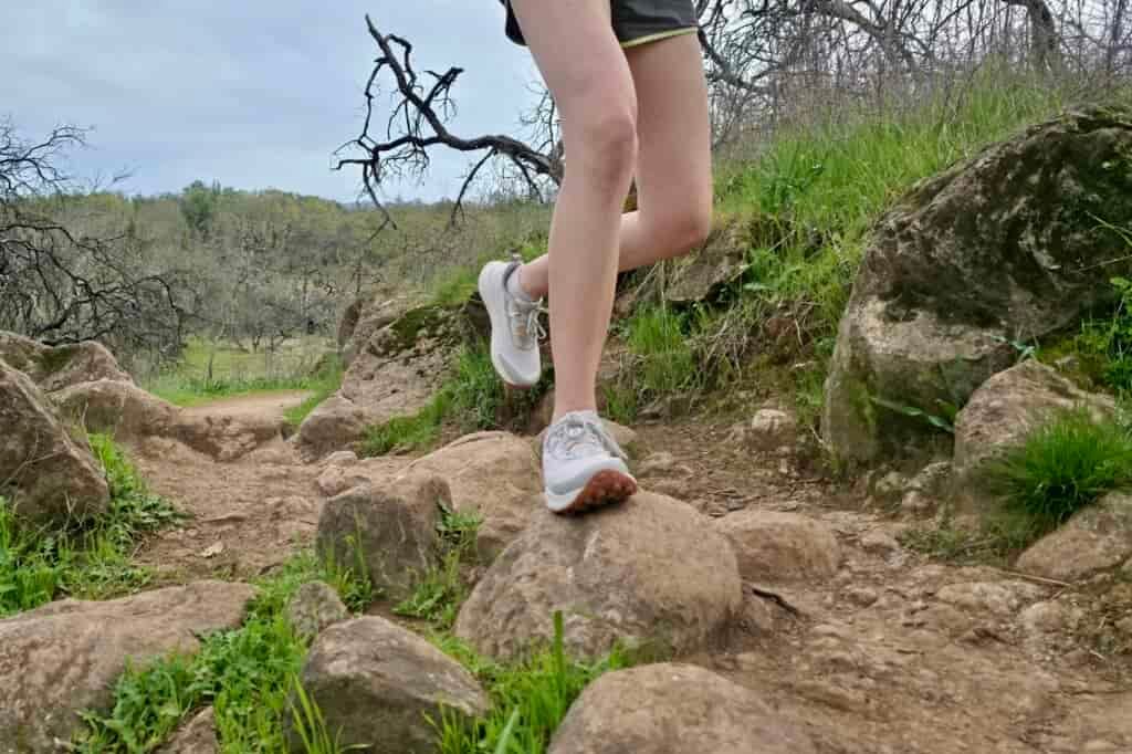 A trail runner runs along a rocky wooded trail in a pair of Saucony shoes.