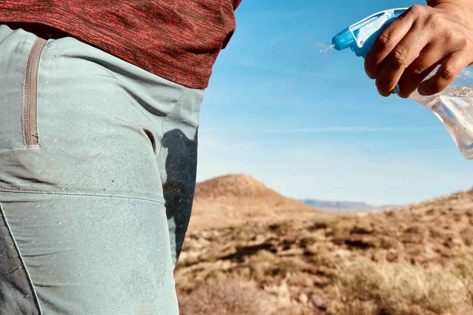 Close up of a hand holding a squirt bottle and spraying the Baleaf pants with water. There is a desert landscape in the background.