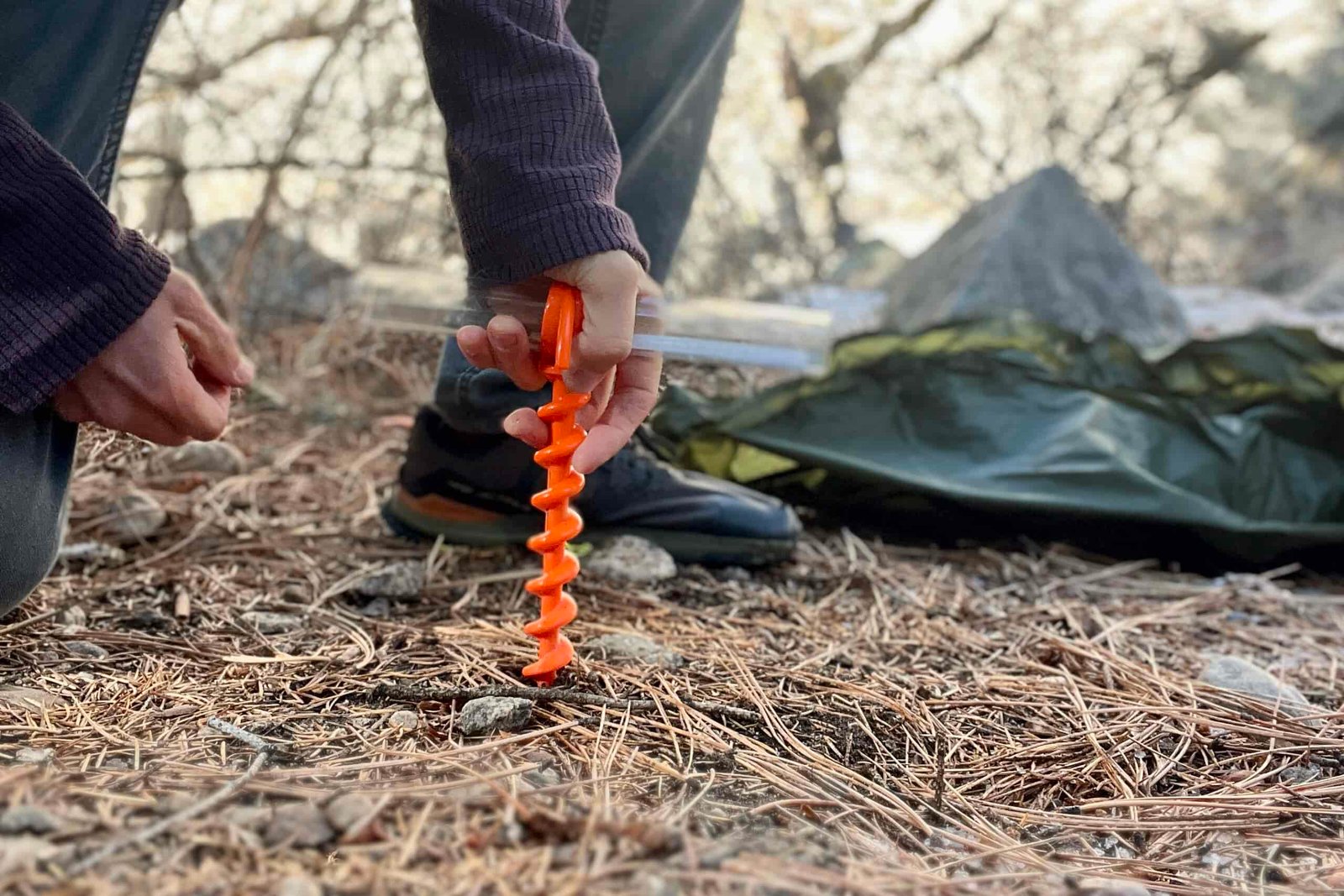 A hiker screws a corkscrew tent stake into piney soil with a tent in the background.