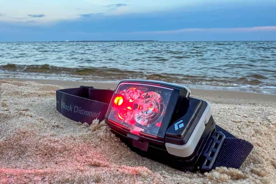 A headlamp sits in the sand near the ocean. The red light is illuminated on the headlamp.
