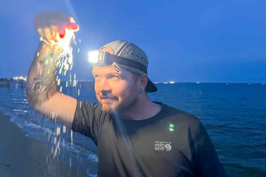 A man sprinkles sand down in front of his headlamp. The ocean is in the background.