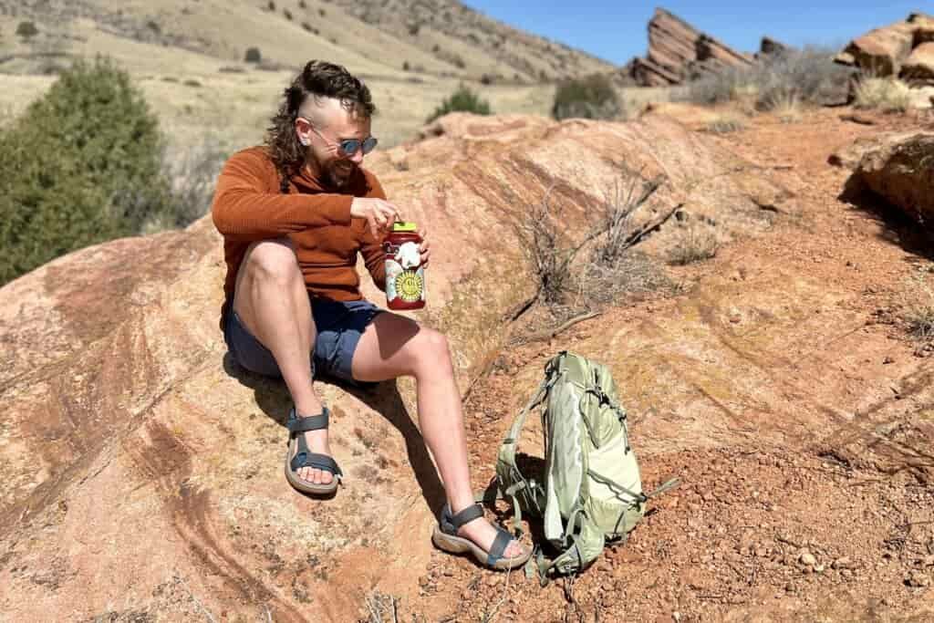 A man smiles as he sits on a rock in a desert setting and opens a water bottle with a backpack at his feet.