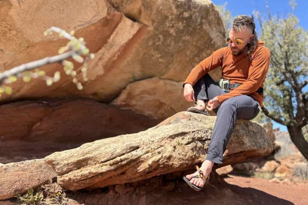 A man in a hoodie and pants sitting on a rock putting on a pair of sandals