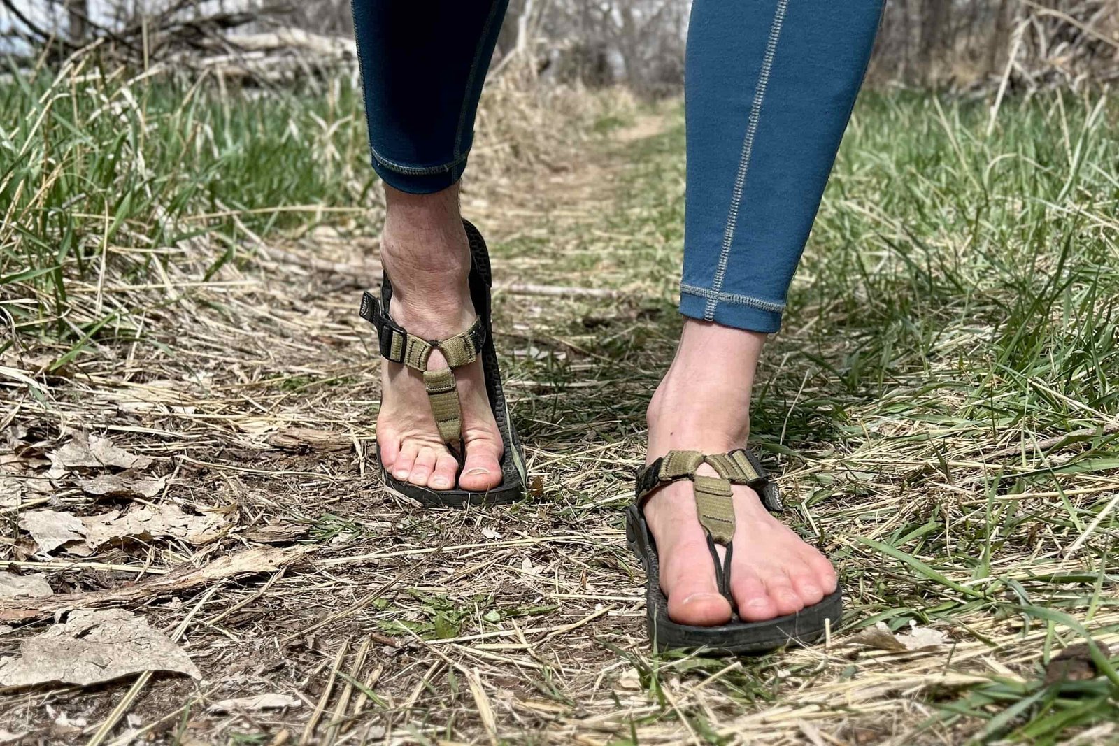 Close up image from the knees down of a person walking in sandals along a grassy path.