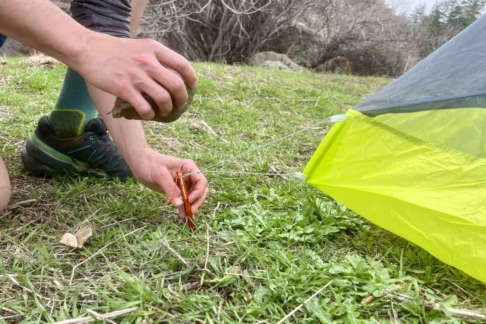 a close up of a hiker pounding in a tent stake with a rock