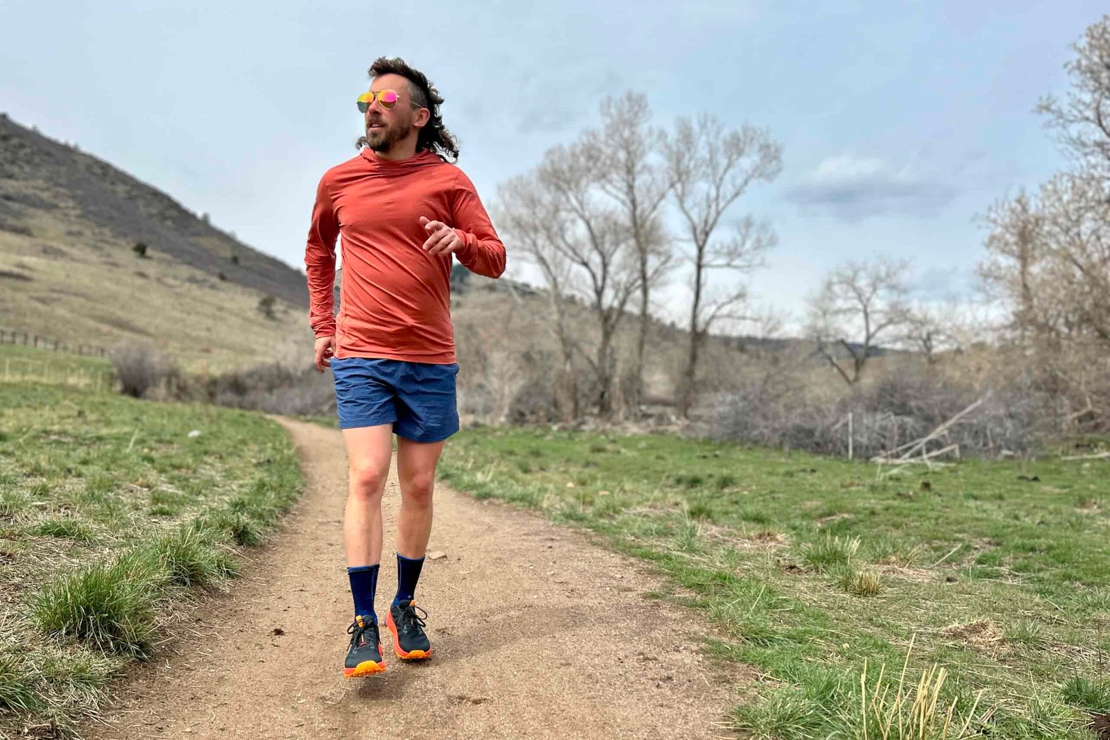A man running down a trail towards the camera wears sunglasses and trail running attire in a hilly landscape.