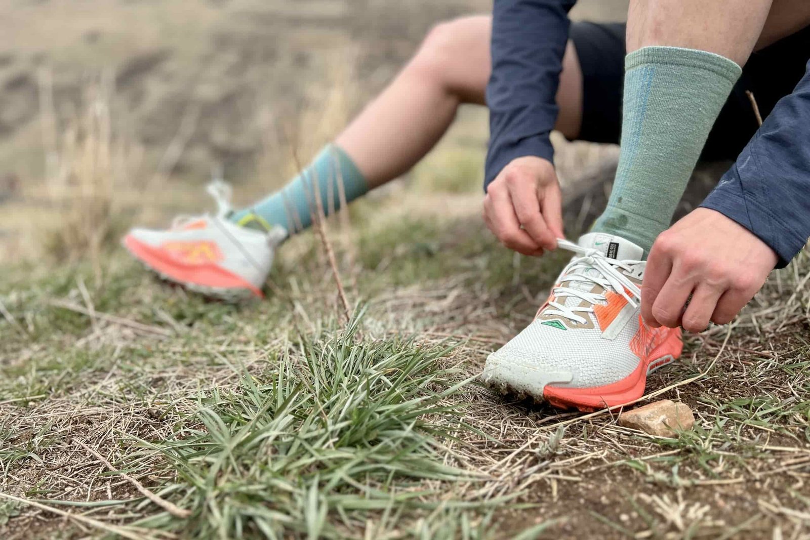 Close up from the waist down of a person tying a pair of shoes as they sit on the ground in a grassy patch.