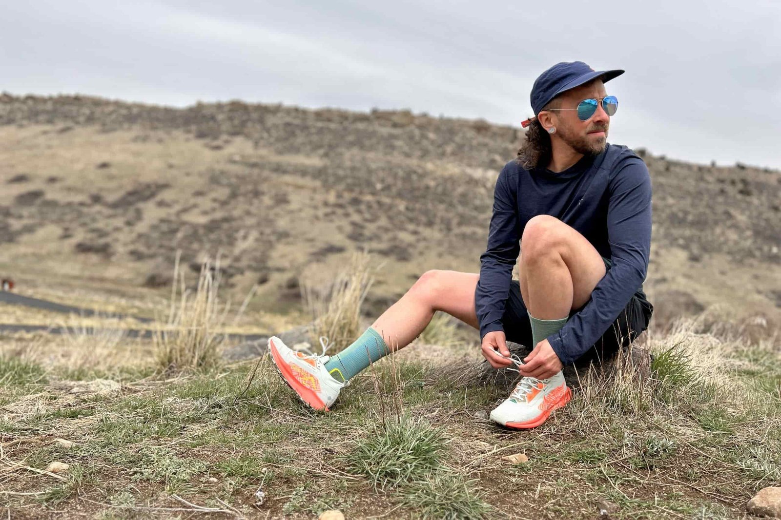 A man in trail running attire and blue sunglasses ties his shoes and looks off in the distance.