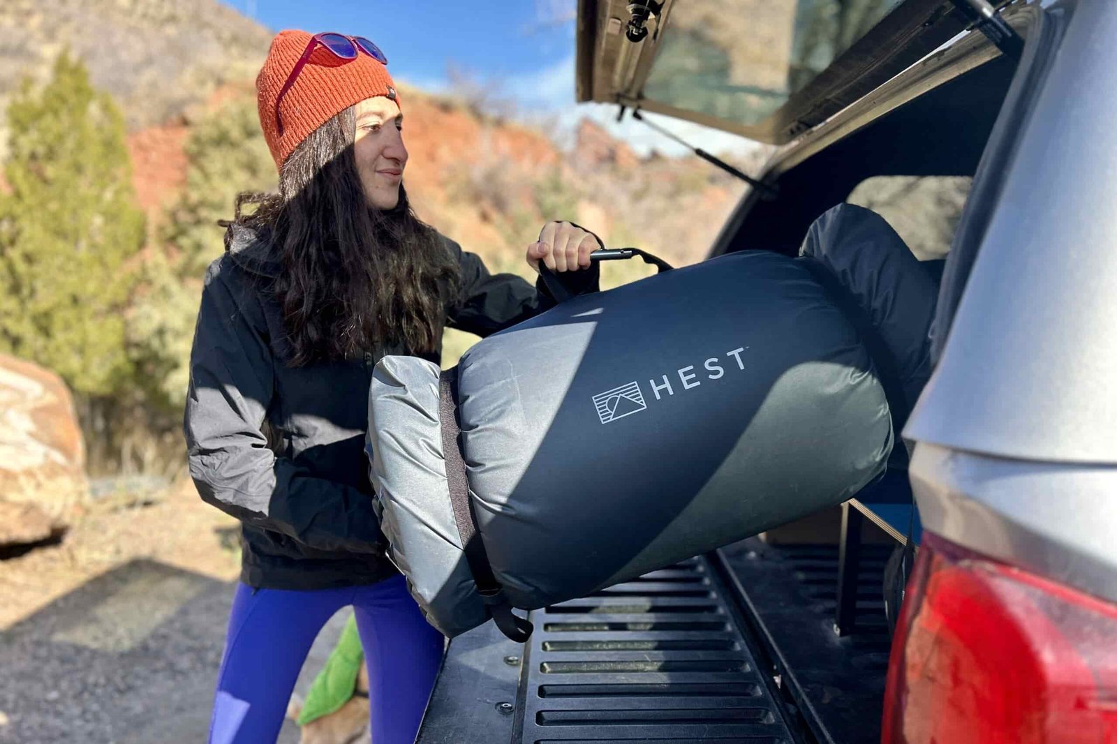 A woman puts a large, rolled-up camping mattress into the back of a pickup truck.