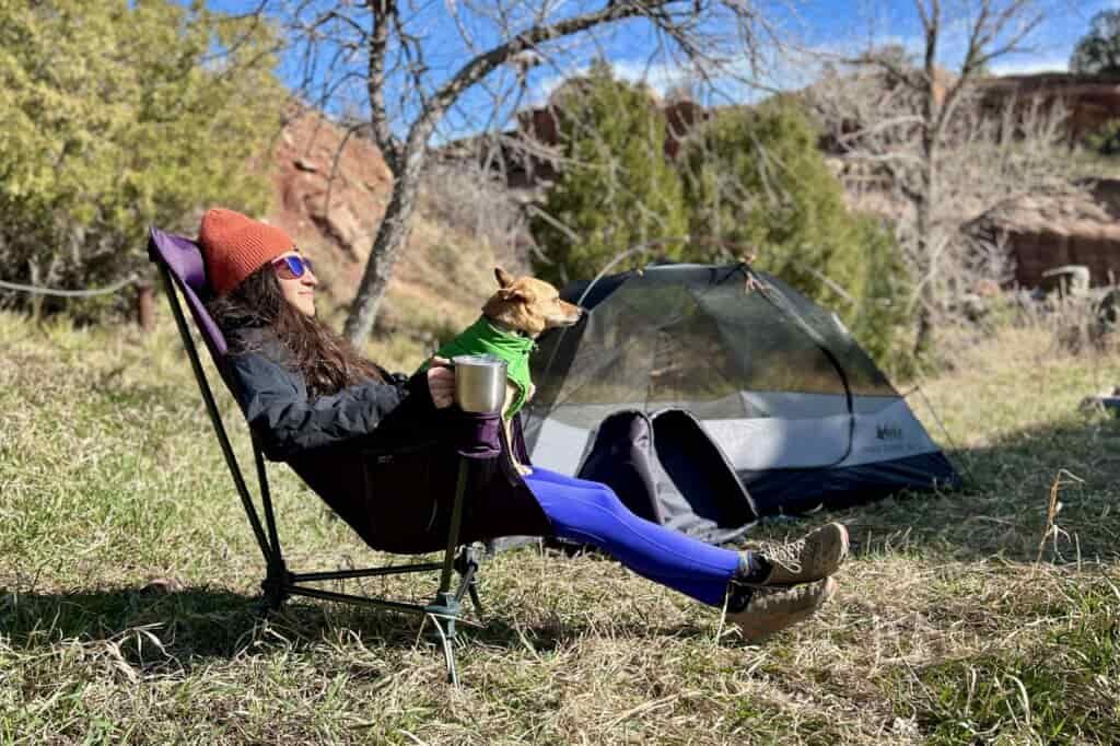 A woman leans back in a camping chair in a forested setting with her hand on a large mug and a small dog on her lap.