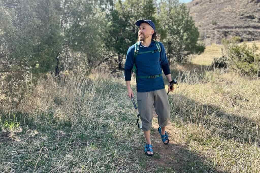 A man hikes through a forested setting in a backpack and sandals.