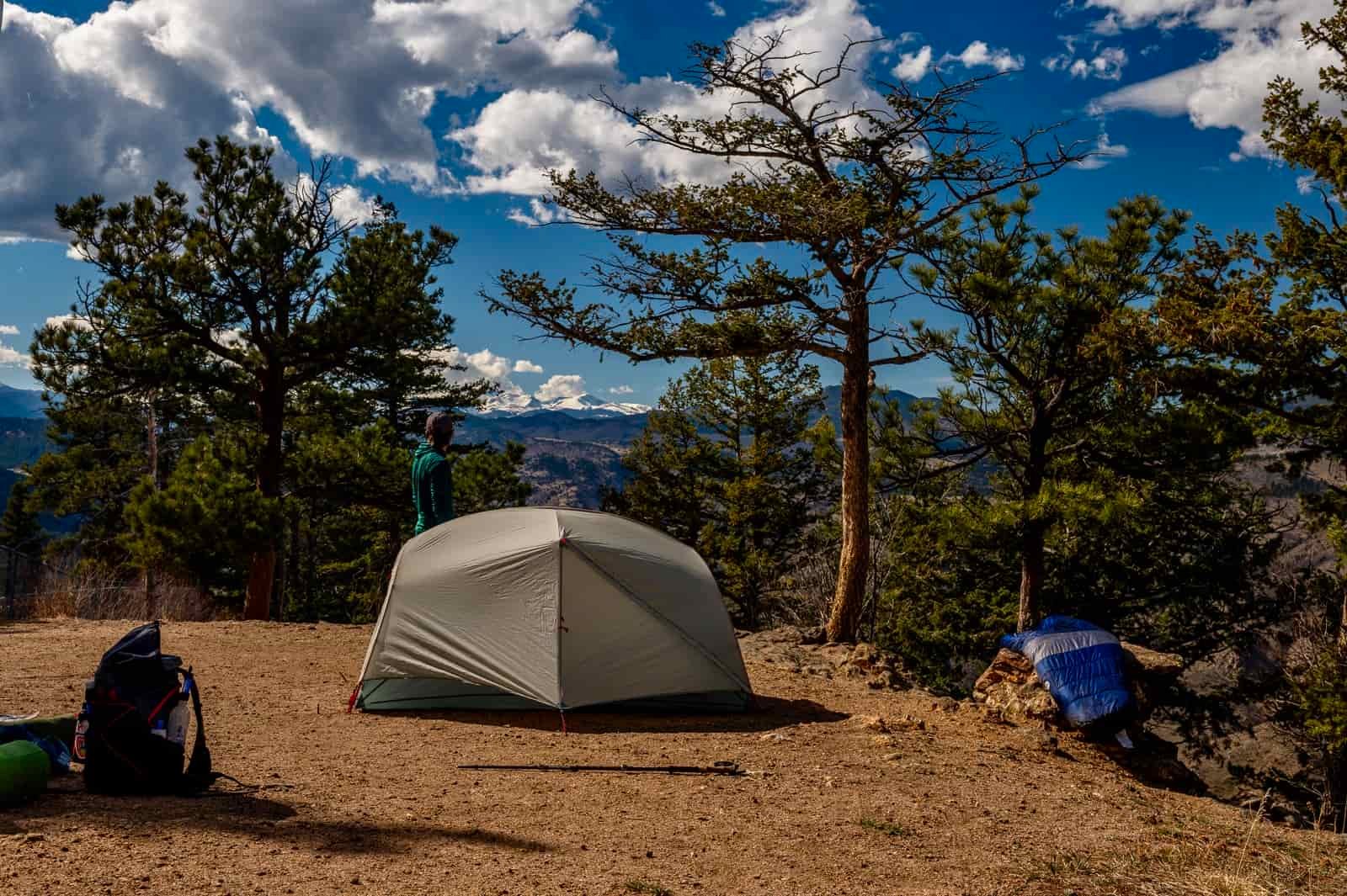 A hiker stands next to a tent looking off into the distance next to an msr tent and other gear sprawled nearby