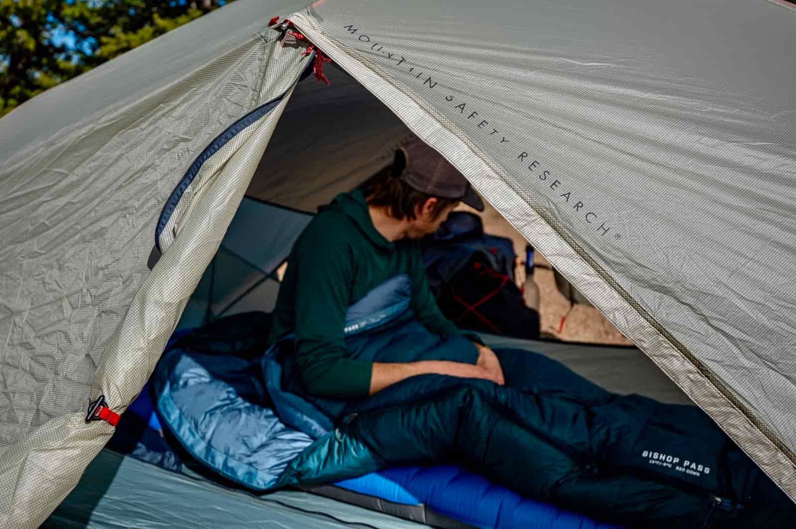 A hiker sits in a ten in a sleeping bag with the tent door in the foreground.