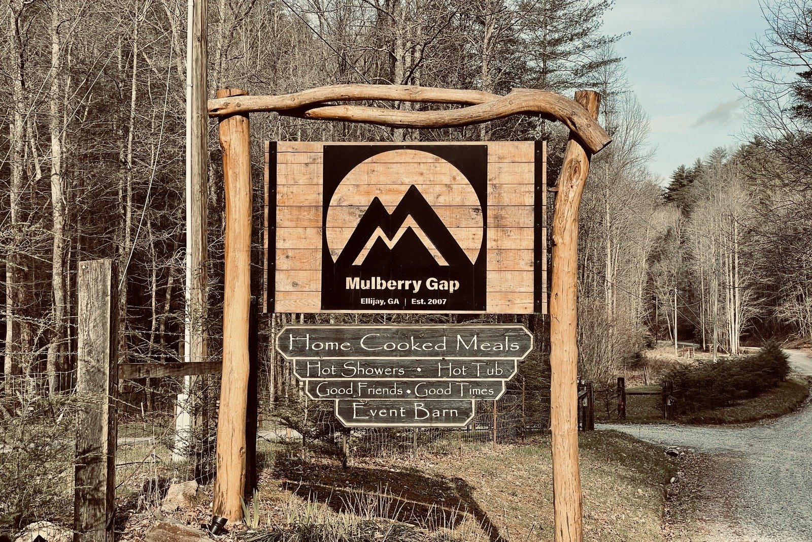 A large, wooden sign for Mulberry Gap Adventure Camp. The camps logo is large and takes up most of the sign. Below it proclaims "home cooked meals, hot showers, hot tub, good friends, good times, event barn". There is a dirt road and forest in the background.