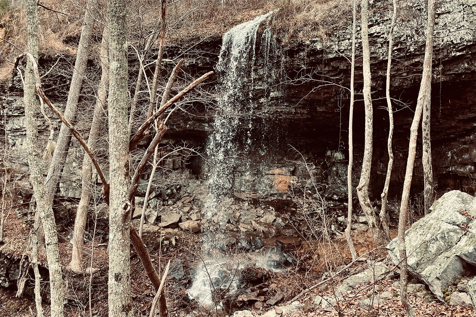 View of a waterfall free falling from a rock ledge through some trees down to more rocks below.