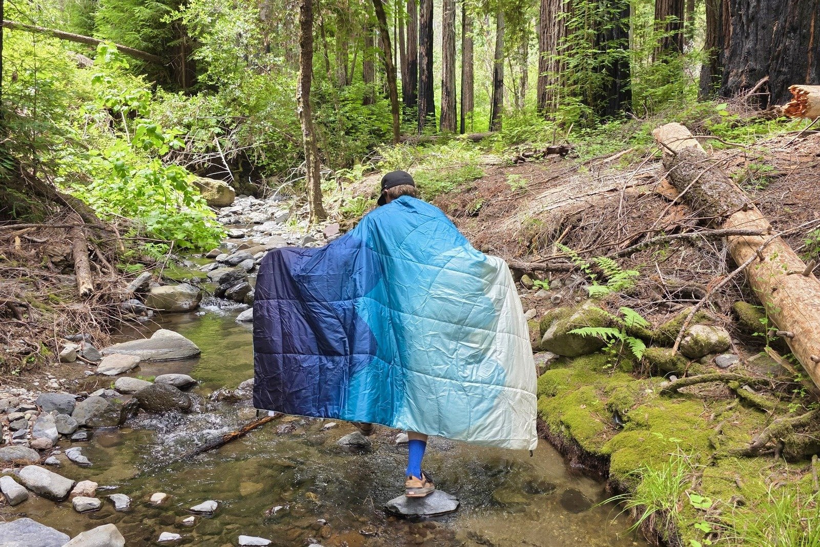 A man walks along a creek while wrapped in a camping blanket.