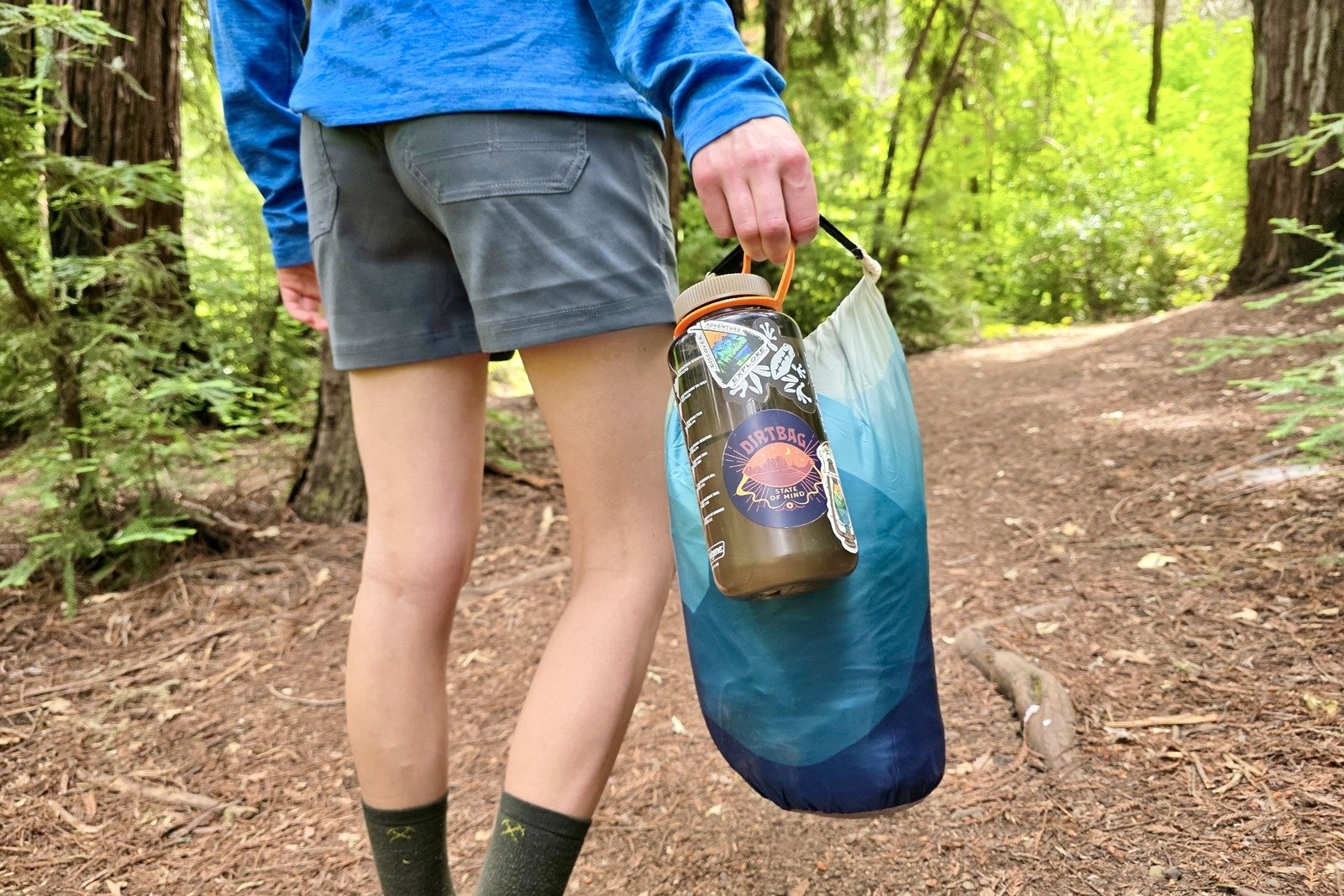 A woman stands holding a blanket in its stuff sack next to a Nalgene to compare size.