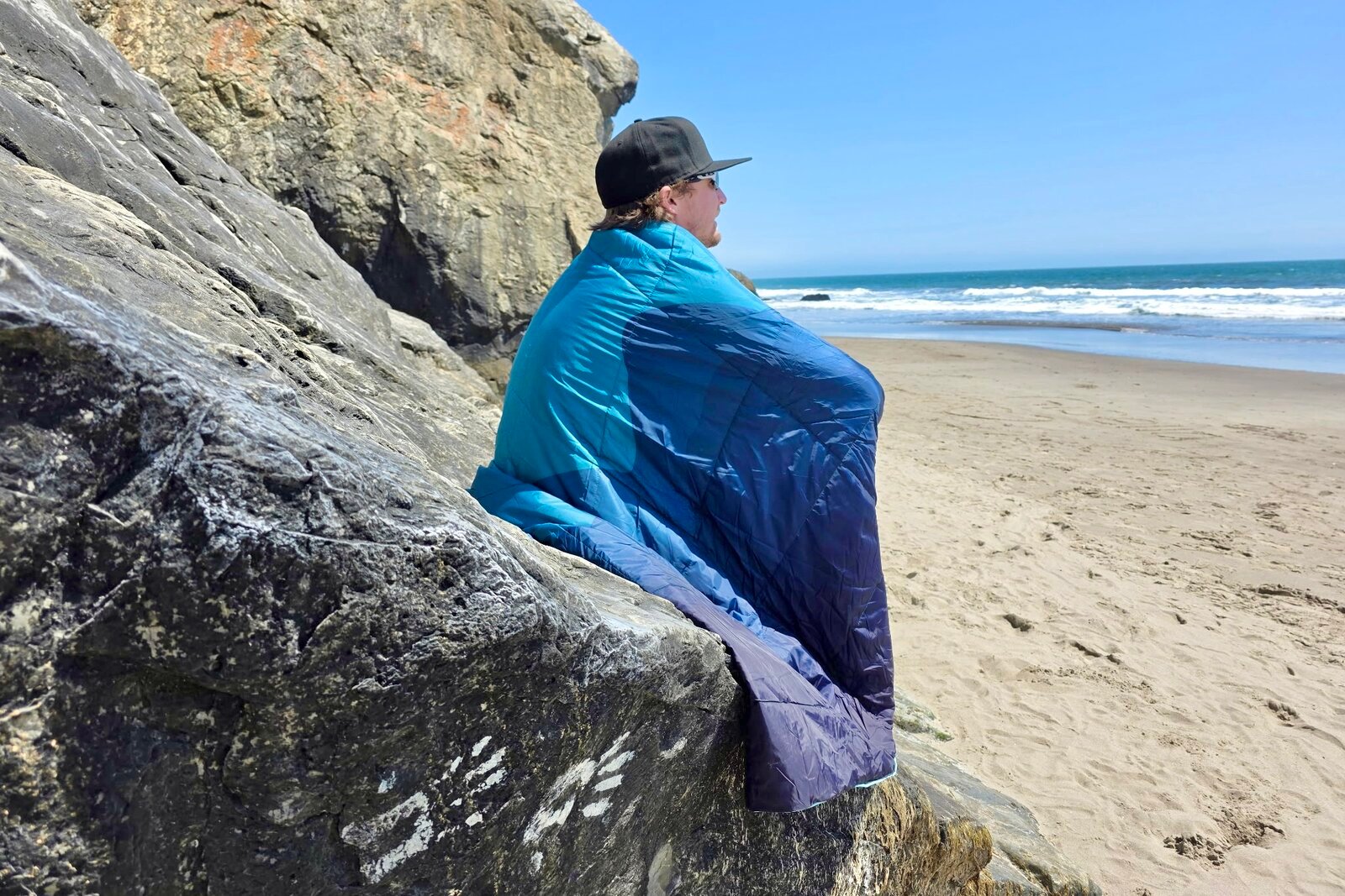 A man sits on a rock overlooking the ocean, while wrapped in a blanket.