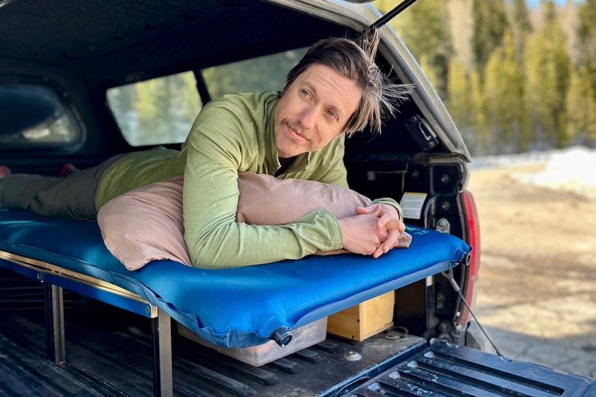 A man in a green shirt lays on a camping mattress in the back of a pickup truck.
