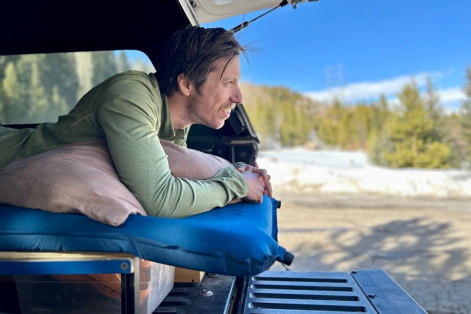 A man in a green shirt looks out at a blue, wintry scene while laying on a mattress in the backof a pickup truck.