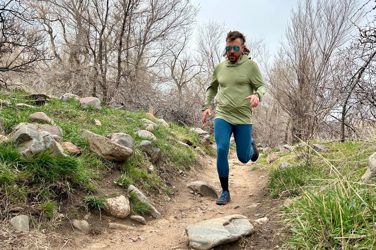 A man wearing a sun shirt, leggings, and aviator sunglasses runs down a trail in a wooded area.