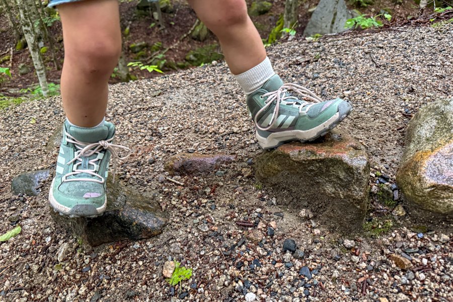 Child hiking over wet rocks on a forest trail while wearing Adidas Terrex AX4R Mid shoes.