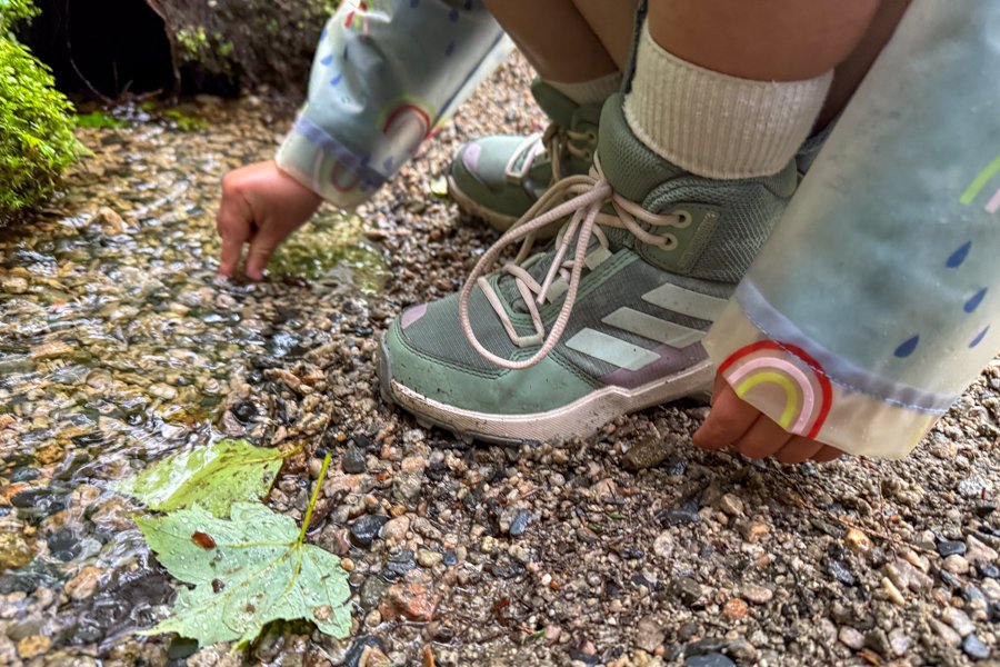 Child crouched at the edge of a shallow creek in Adidas Terrex AX4R Mid shoes and a colorful raincoat.