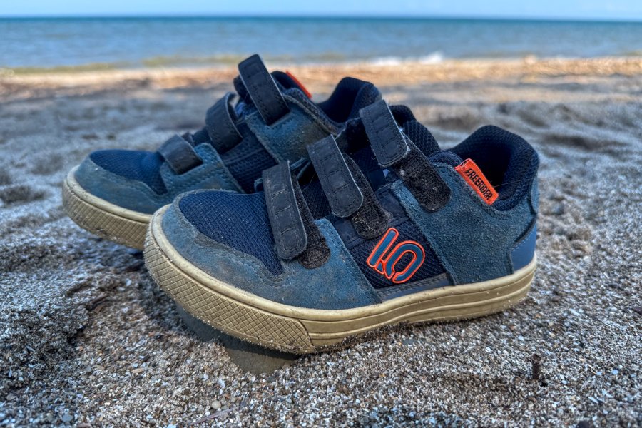 Close-up of worn Five Ten Freerider kids shoes sitting on sandy ground near a lake.