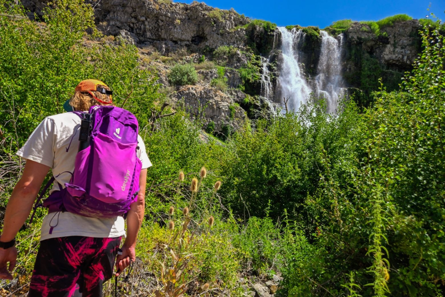A person wearing a purple Deuter Speedlite Pro daypack hikes toward a large waterfall in the background.