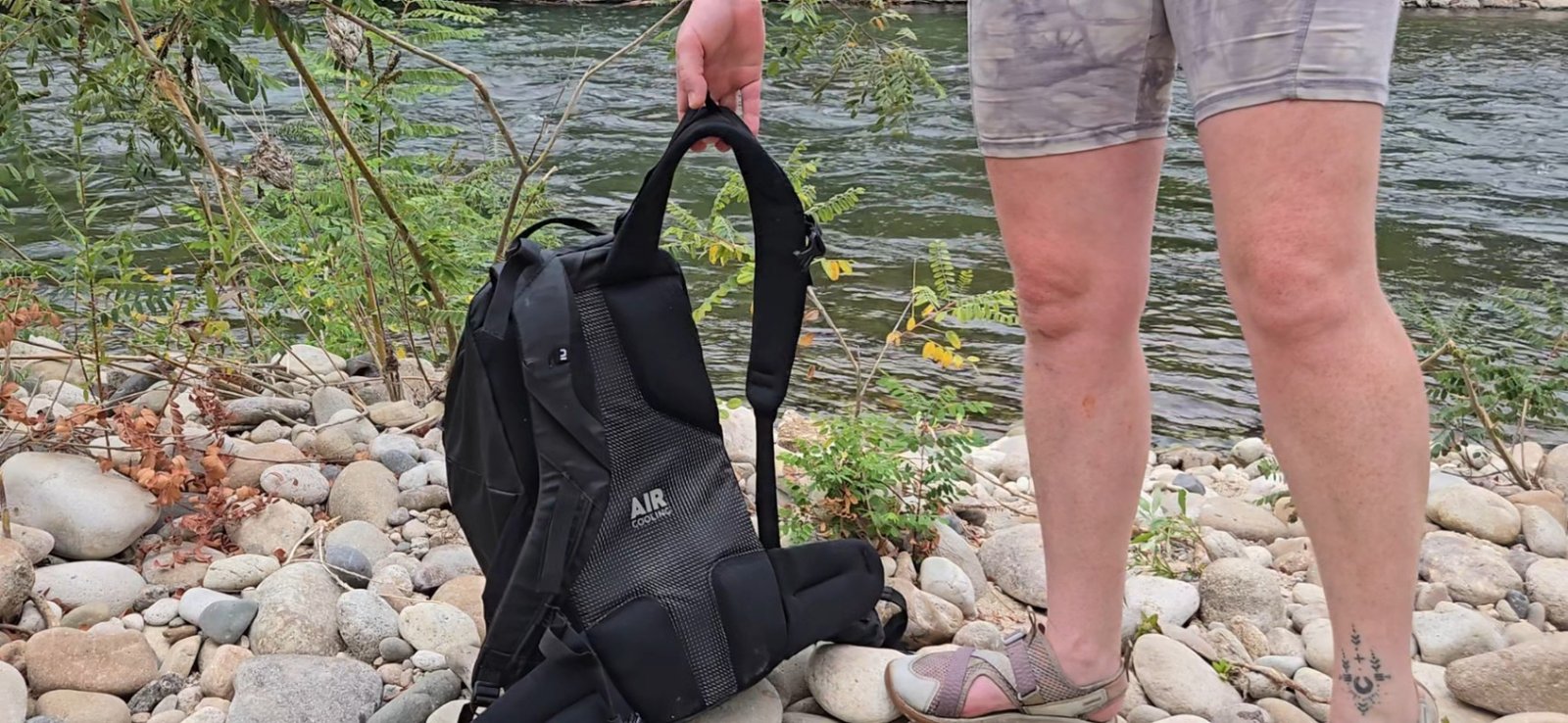 A person stands on a cobble riverbank and holds the Decathlln Quechua MH100 20L pack before they lift it to put it on. You can see the ample back padding and "Air Cooling" ventilation.