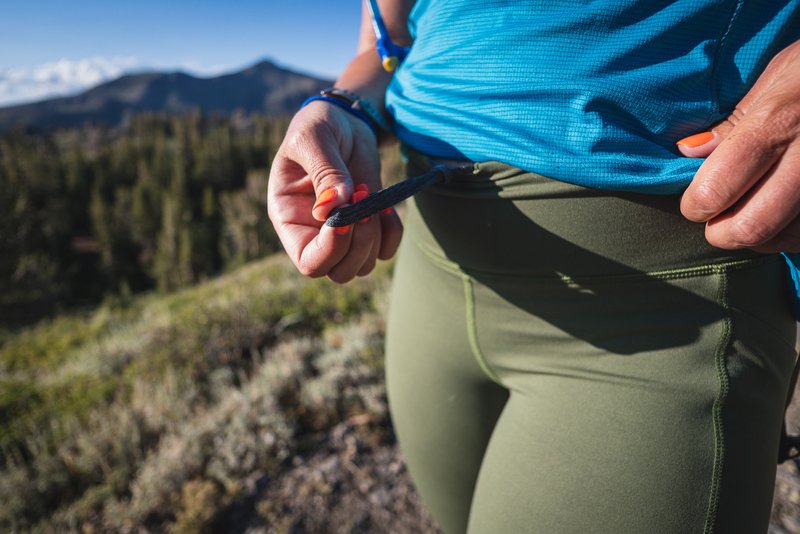 Women in green hiking leggings showing the infinity drawstring, wearing a bright blue shirt, with mountains and trees in the background.