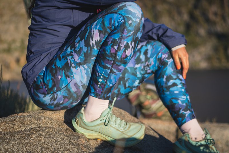 Woman wearing bright blue leggings in a camouflage like pattern on a hike. She's sitting on a rock with knees bent and tucked in tight to her body. She's wearing light teal trail runners.