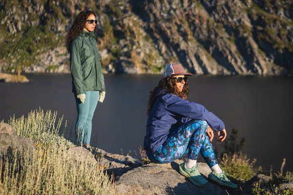Woman wearing bright blue leggings in a camouflage like pattern with a purple rain jacket, sunglasses and a hat. She's sitting in the foreground, with knees bent and arms resting on her knees. Her friends is standing behind her, closer to the lake in the background. She's wearing lighter blue leggings, no pattern, with a green rain jacket, and sunglasses. Both have curly hair.