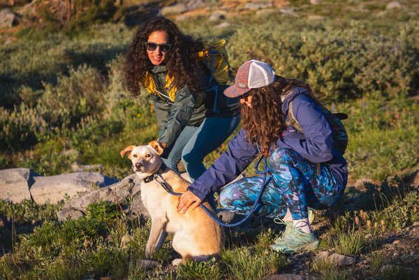 Woman wearing bright blue leggings in a camouflage like pattern on a hike with her friend and her dog. She is crouched down petting the dog. Her friend (in light blue leggings and a green jacket) is bent slightly at the waist also petting the dog.