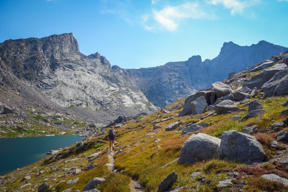 a trail cut in on an alpine meadow hillside with a glittery lake below and granite towers in the background