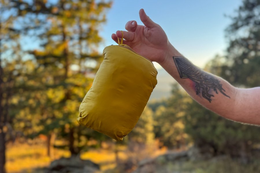 A hand holding the packed Cotopaxi Fuego Hooded Down Jacket in its stuff sack, with trees and sunset colors in the background.