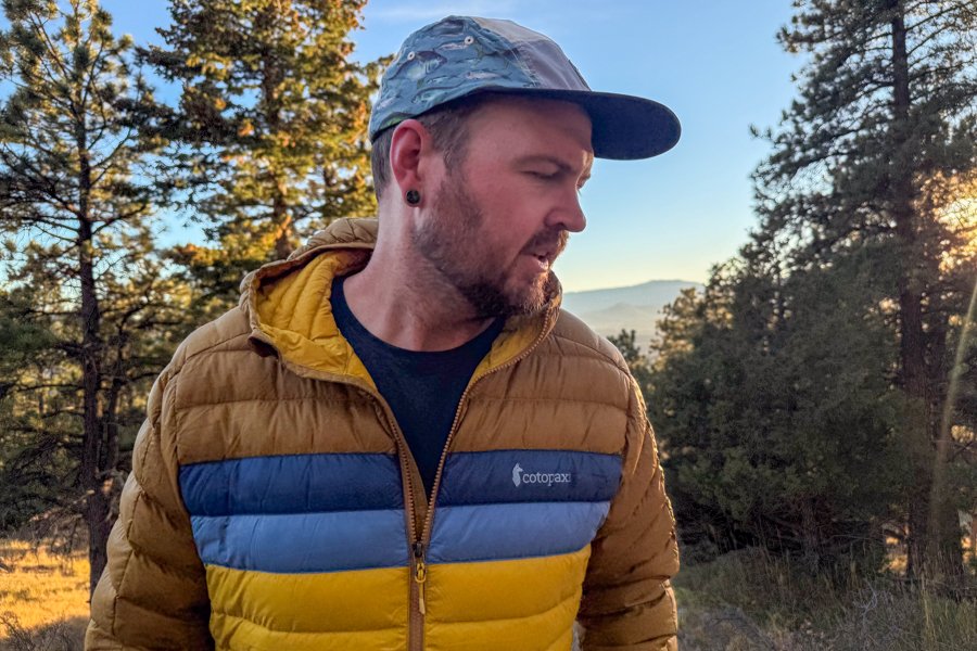 A man wearing a Cotopaxi Fuego Hooded Down Jacket in the mountains at sunrise, looking down as warm morning light filters through pine trees.