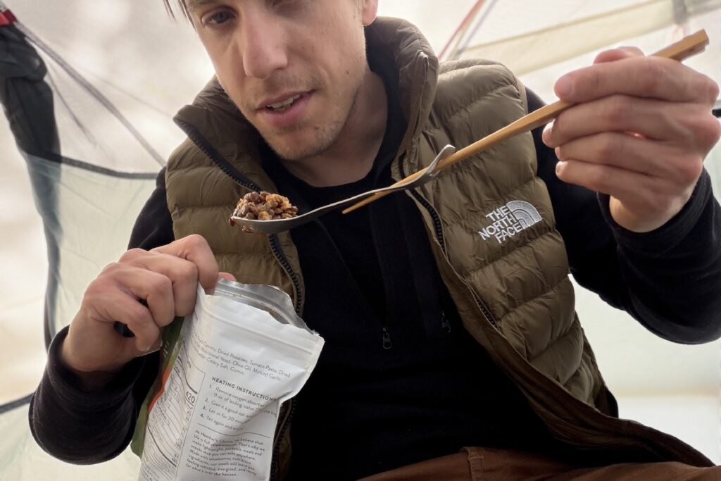 a hiker scoops a bite of food from a meal pouch with a spork extended with chopsticks