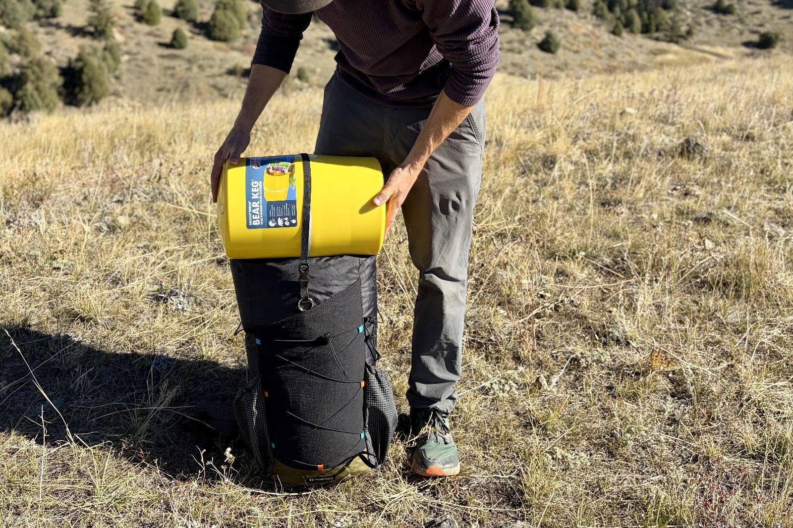 a hiker holds a backpack on the ground with a bear canister on top
