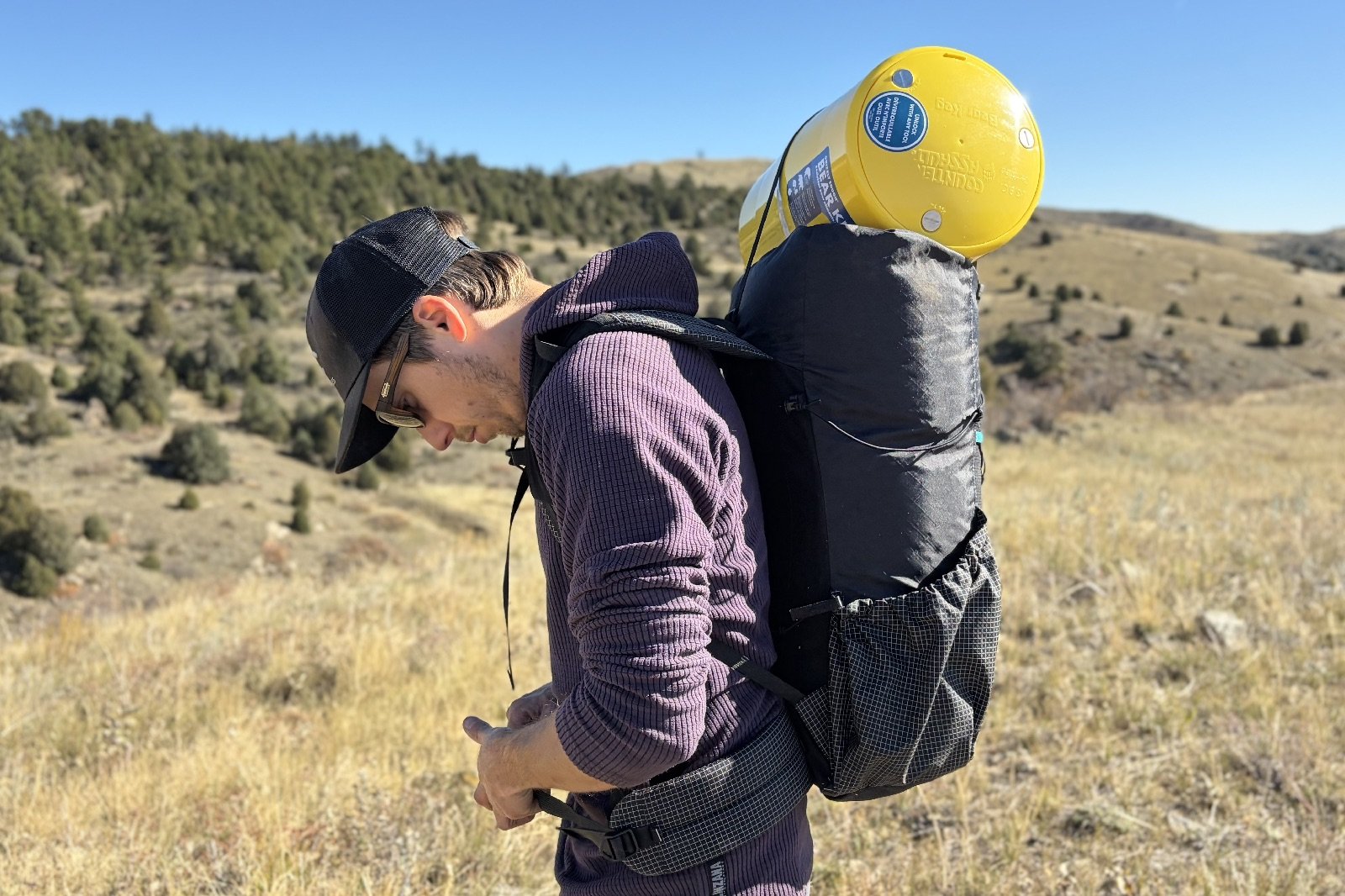 a hiker buckles the waist belt on his backpack with a yellow bear canister strapped on top